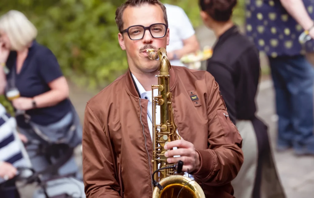 Ein Mann, der Saxophon sspielt und eine braune Jacke trägt. Im Hintergrund läufen Menschen vorbei.