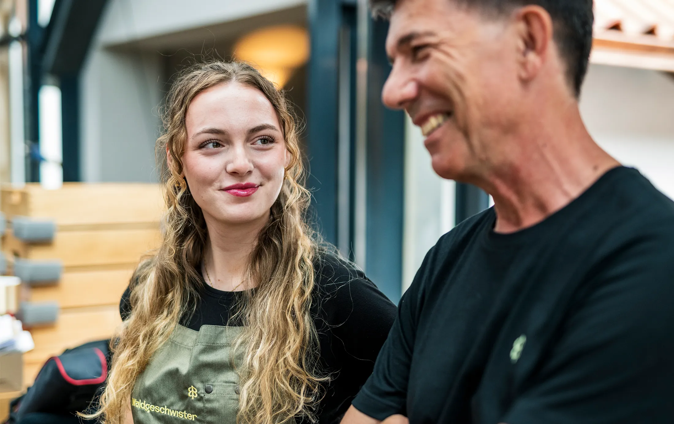 Eine Angestellte mit blonden Locken und einer Schürze und ein Mann mit schwarzem T-Shirt