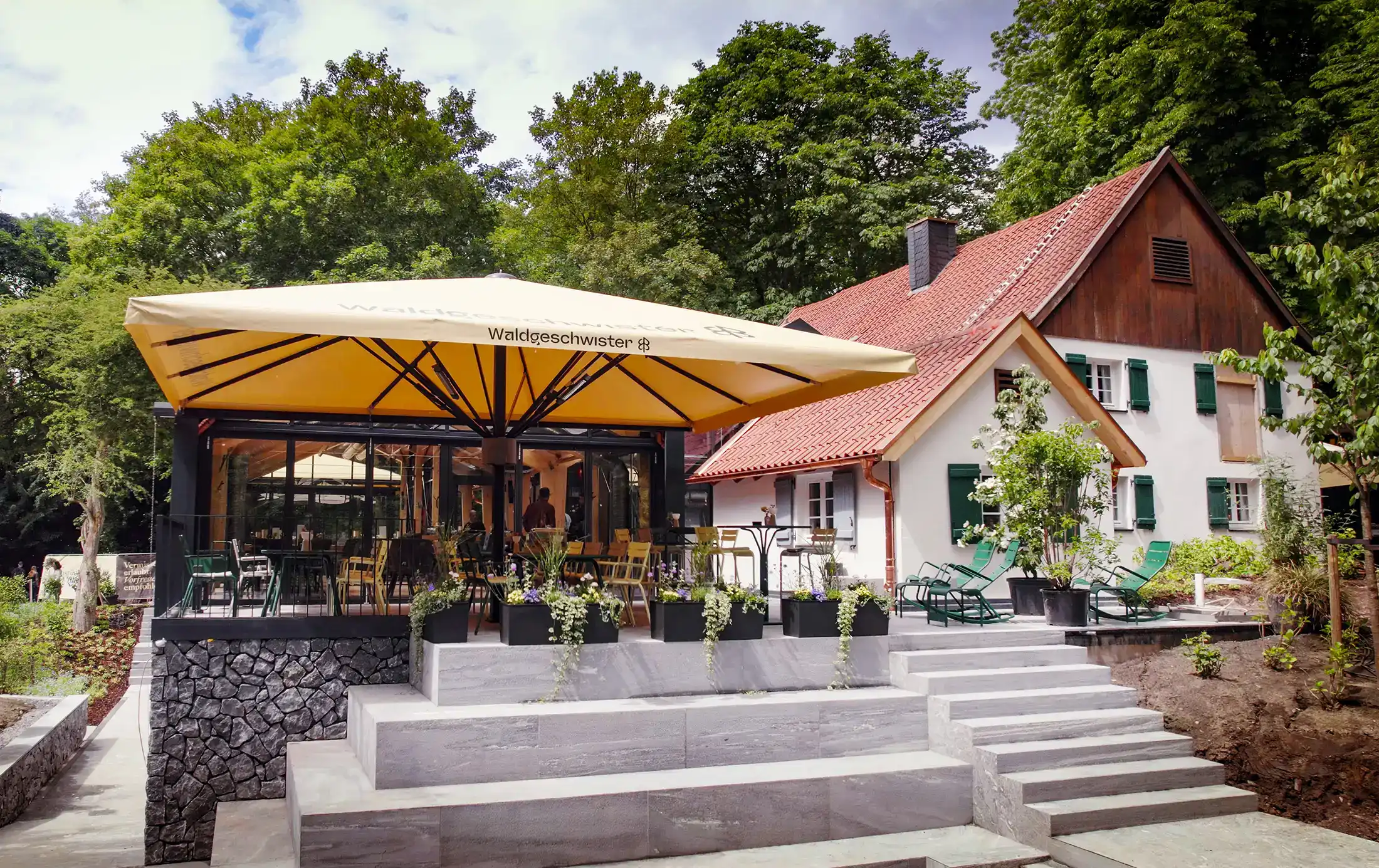 Die Terasse des Forthauses mit einem gelben Sonnenschirm über Sitzgelegenheiten und im Hintergrund das Forsthaus