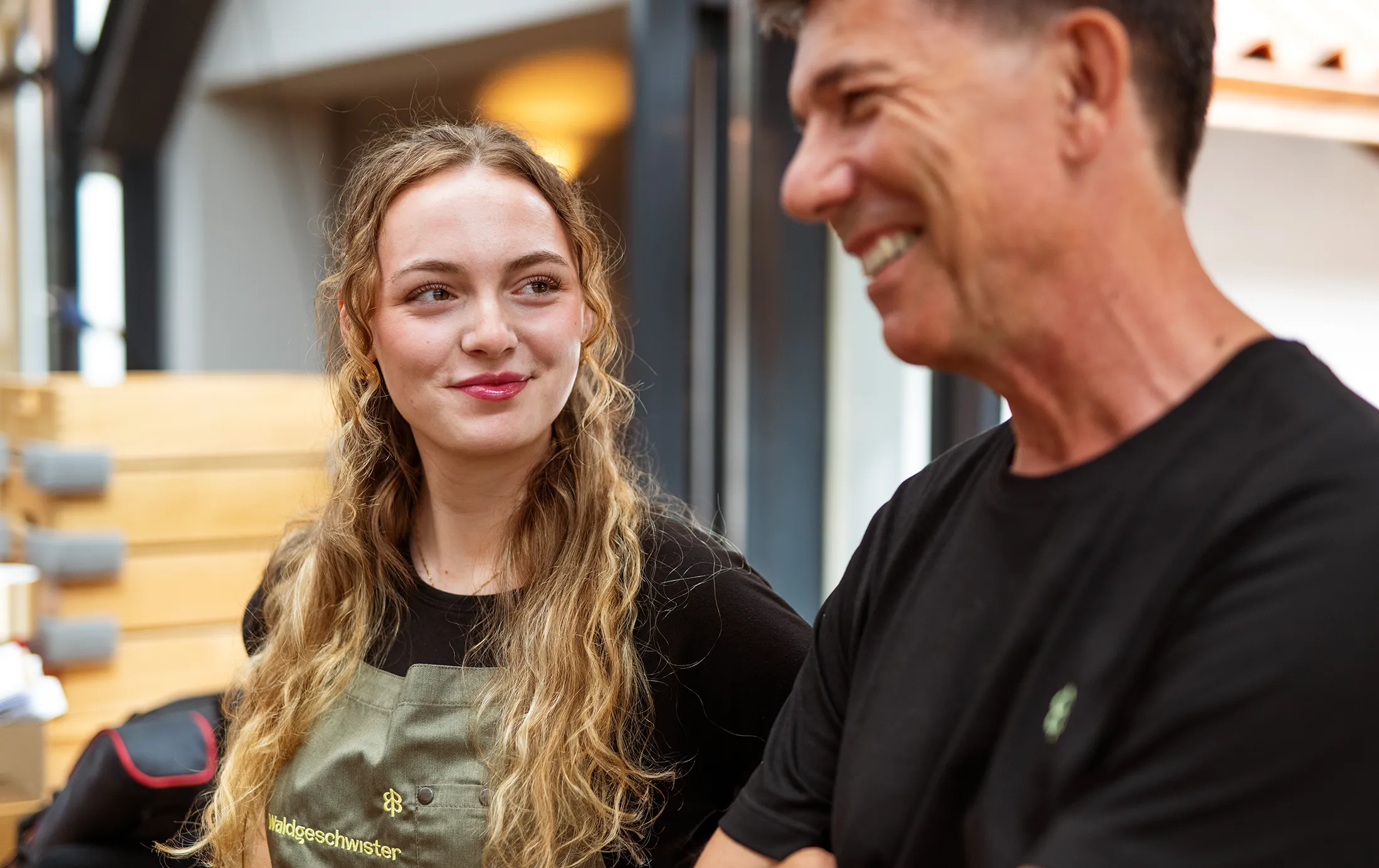 Eine Angestellte mit blonden Locken und einer Schürze und ein Mann mit schwarzem T-Shirt